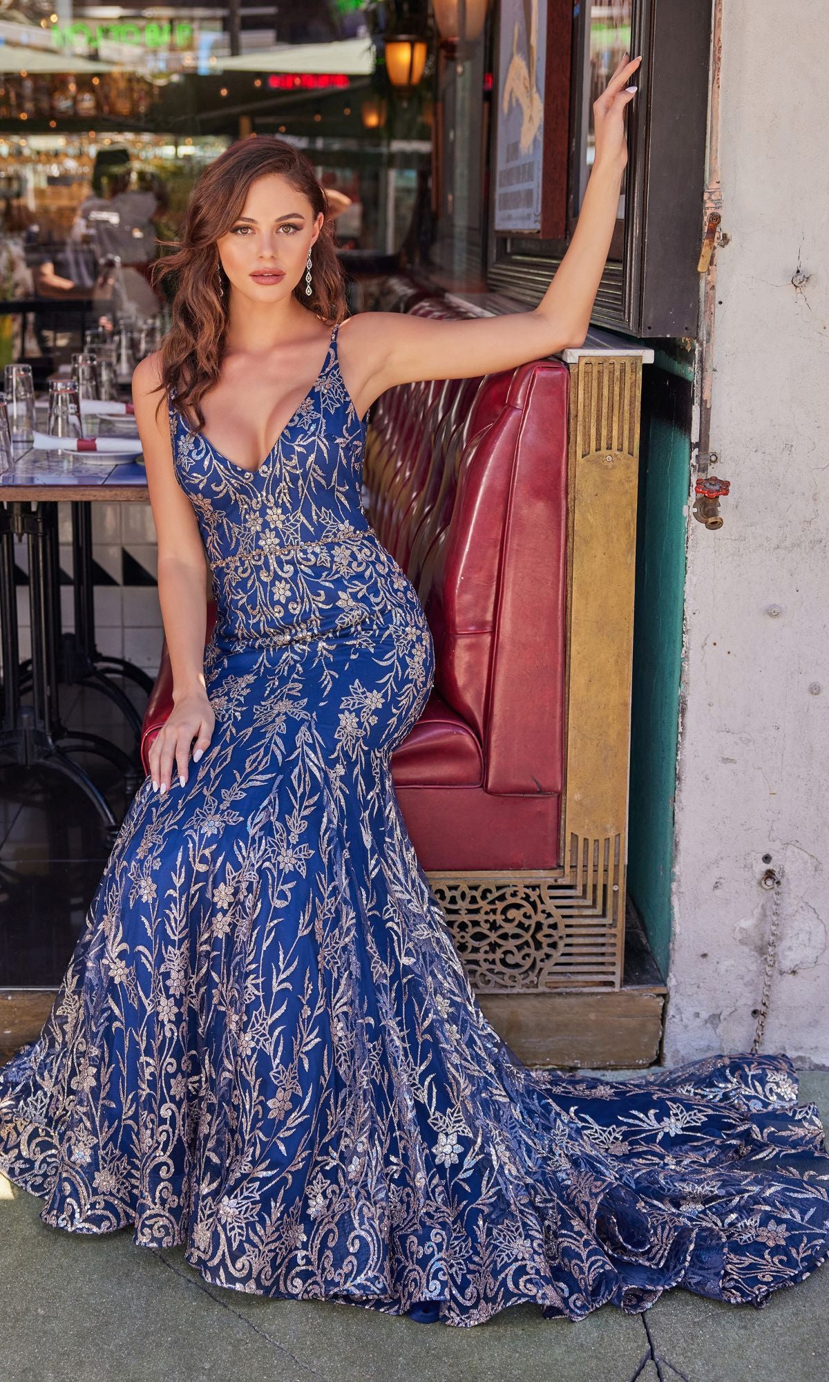 Wearing the Colette CL2021 Long Mermaid Glitter Prom Dress in blue and gold, a woman sits elegantly on a red restaurant booth, one arm gracefully resting on the back.